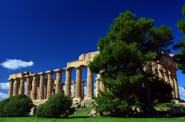 Rovine di un antico tempio con colonne doriche, circondate da alberi sotto un cielo blu.