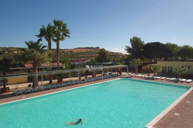 Ampia piscina all'aperto con lettini, circondata da palme e edifici, sotto un cielo soleggiato.
