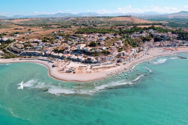 Vista aerea di una costa con mare turchese, spiaggia e case in lontananza.