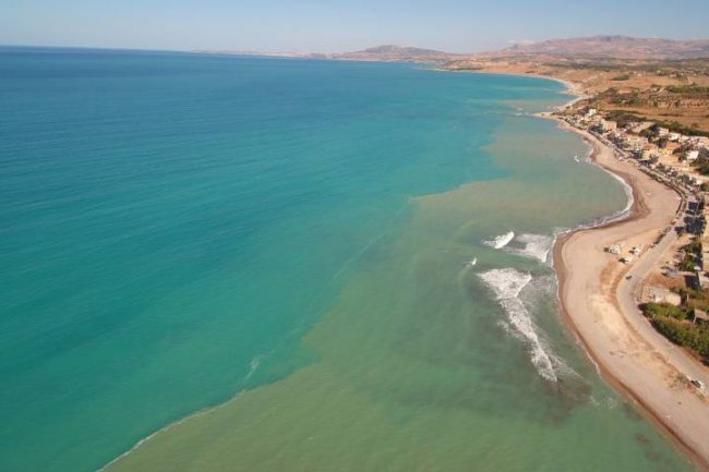 Panorama costiero con mare blu e spiaggia, strade e colline visibili sulla destra.