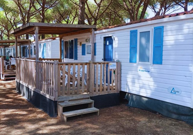 Casa mobile bianca con persiane blu e veranda in legno tra gli alberi.