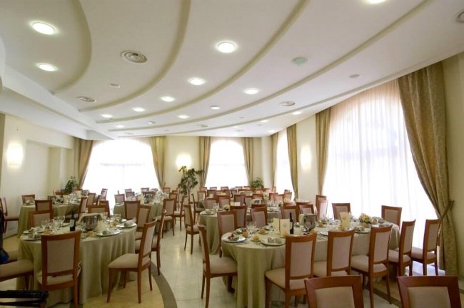 Sala ricevimenti elegante con tavoli rotondi apparecchiati, finestre grandi e soffitto ondulato.