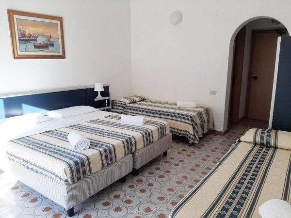 Camera d'albergo con quattro letti singoli, arredamento semplice, e pavimento piastrellato.