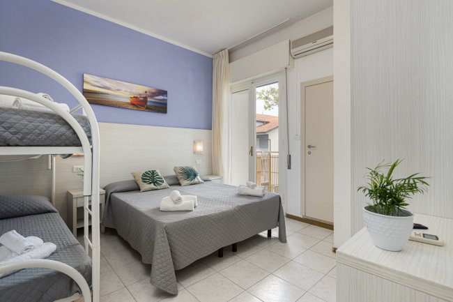 Una camera d'albergo con letto matrimoniale, letti a castello e piante. Parete blu e porta finestra.