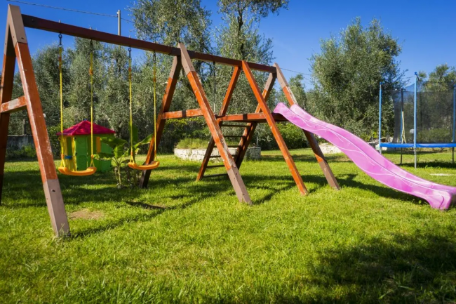 Parco giochi con altalene, scivolo rosa e casetta colorata su erba verde.