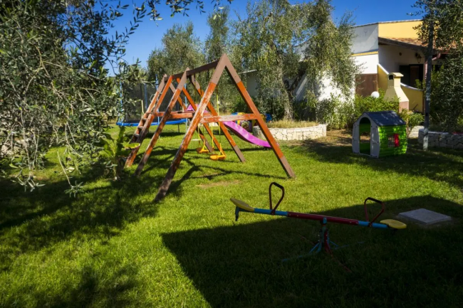 Parco giochi con altalene, scivolo, bilico e casetta, circondato da alberi.