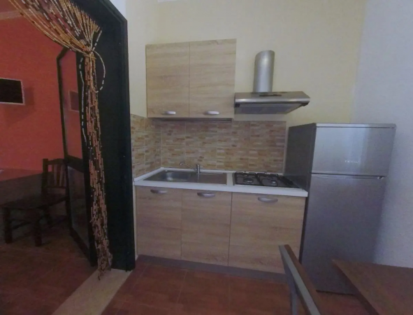 Cucina compatta con mobili in legno, lavello, fornelli, cappa e frigorifero in acciaio.