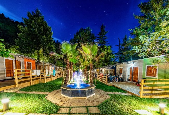 Un giardino notturno con fontana luminosa e bungalow illuminati, circondato da alberi.