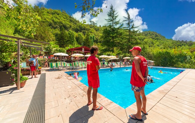 Piscina all'aperto con bagnini in uniforme rossa e ombrelloni verdi, circondata da alberi.