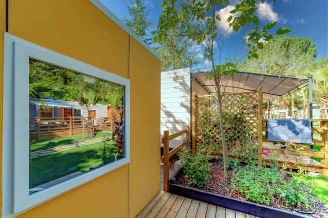 Giardino con terrazza in legno, piante e strutture moderne sotto un cielo azzurro.