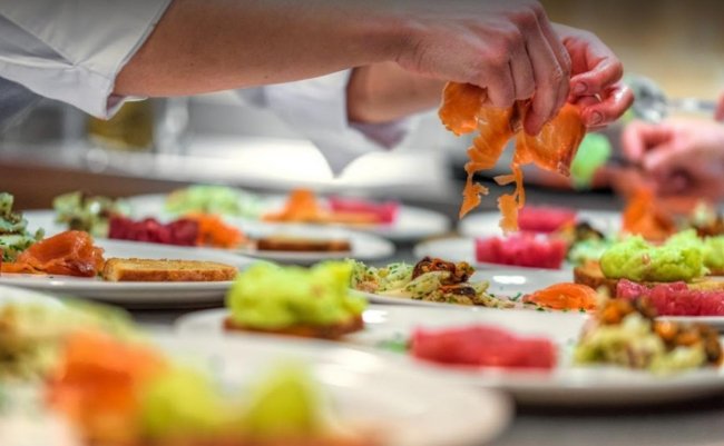 Mani di chef impiattano salmone e verdure su piatti bianchi.