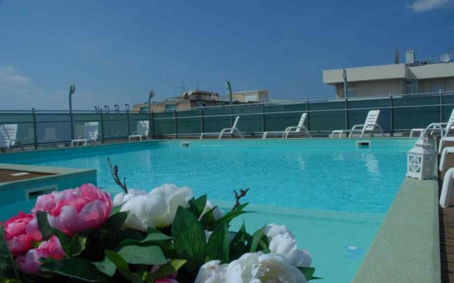 Piscina con lettini bianchi su un terrazzo, fiori in primo piano.