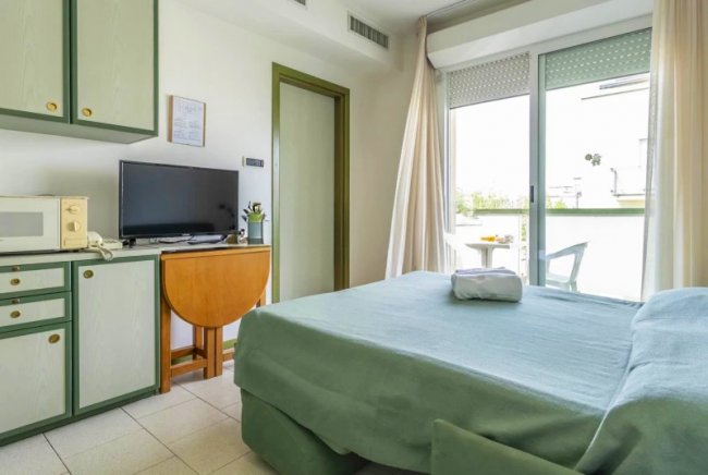 Camera con letto verde, TV su mobile, finestra con tende e balcone.