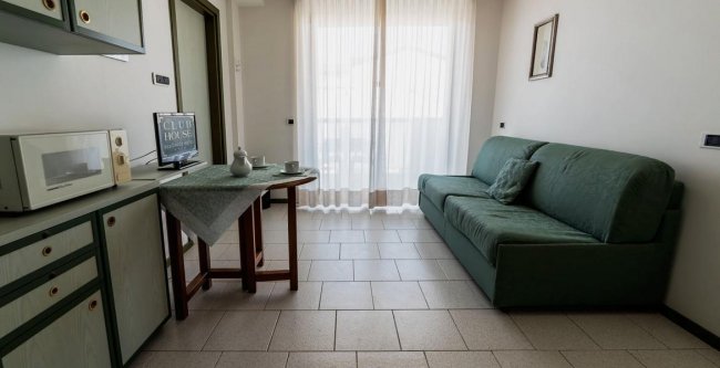 Mini appartamento con divano verde, tavolo con tenda, microonde e grande finestra con tenda bianca.