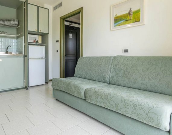 Soggiorno con divano verde chiaro, cucina e porta d'ingresso.