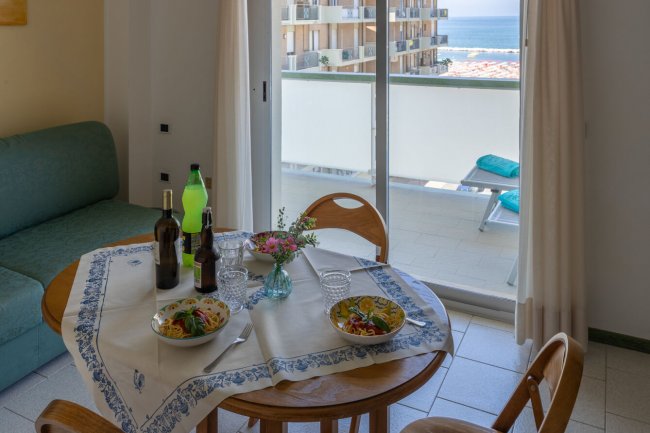 Tavolo rotondo con piatti di pasta e bevande davanti a un balcone con vista mare.