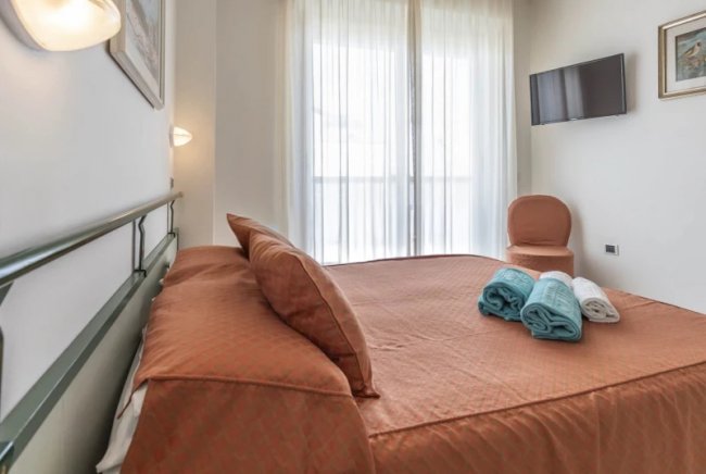 Letto matrimoniale con asciugamani arrotolati, TV a parete e sedia vicino a una finestra ampia.