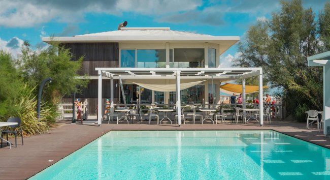 Piscina con vista su un edificio moderno circondato da vegetazione e cielo blu.