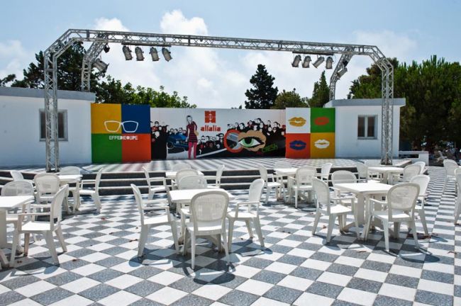 Piazzale con tavoli, sedie e palco colorato.