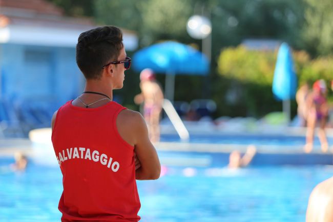 Bagnino di spalle in maglia rossa osserva la piscina.