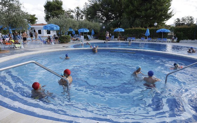 Persone nuotano in piscina circondata da ombrelloni blu.
