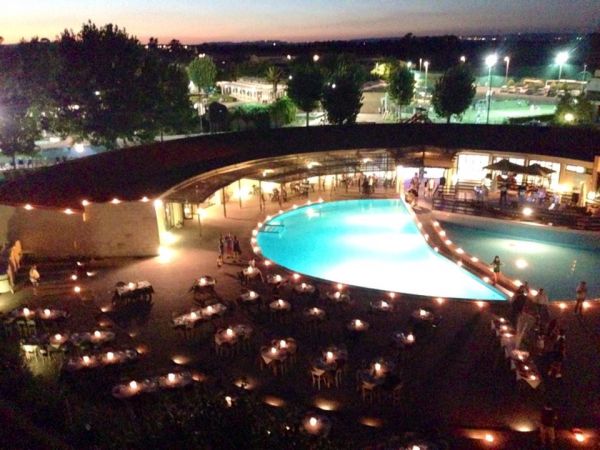 Piscina illuminata di sera, tavoli apparecchiati intorno.