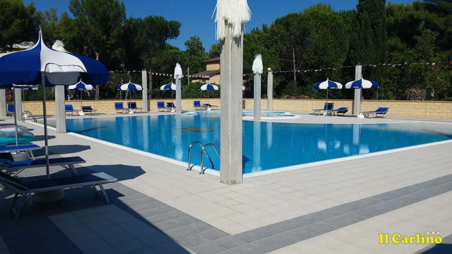 Piscina all'aperto con ombrelloni blu e lettini, circondata da alberi.