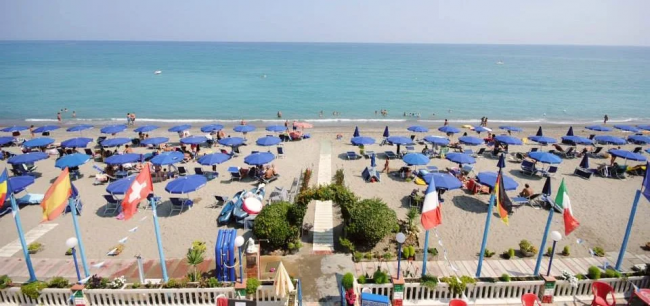 Spiaggia con ombrelloni blu e bagnanti, mare calmo e bandiere sventolanti in primo piano.