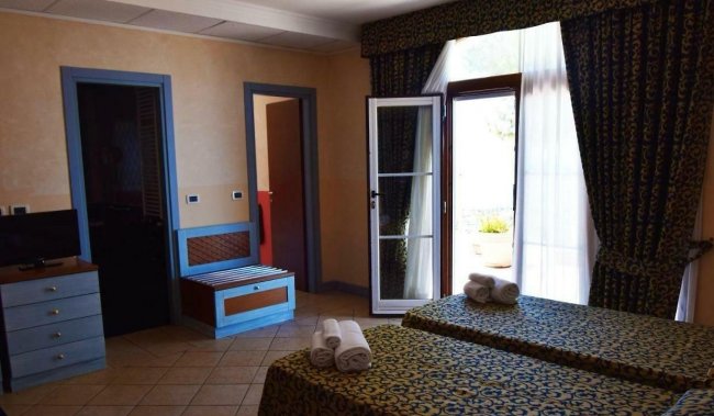 Una camera d'albergo con due letti, asciugamani, un'ampia finestra aperta e mobili blu e legno.