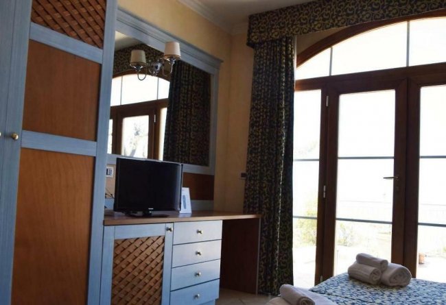 Interno di una camera con letto, asciugamani, scrivania con TV e grandi porte-finestre.