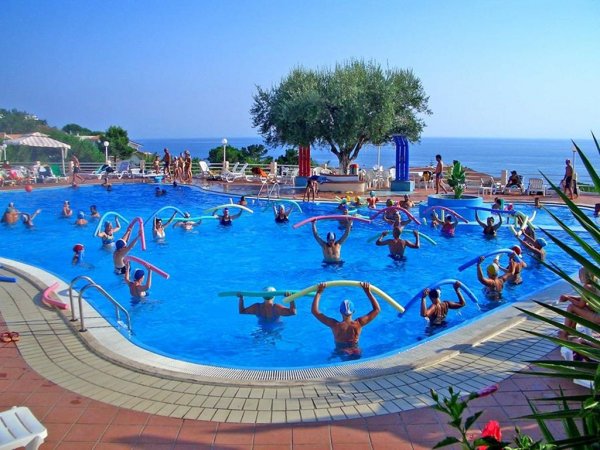 Persone in piscina fanno esercizi con tubi colorati, vista mare e alberi sullo sfondo.