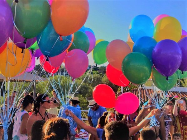 Gruppo di persone tiene palloncini colorati, cielo blu e vegetazione sullo sfondo.