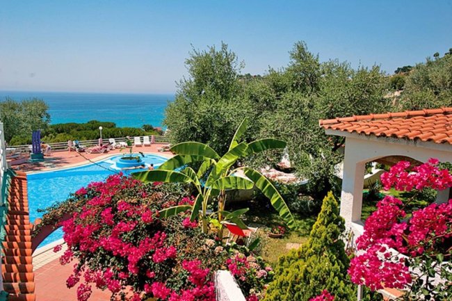Vista di piscina circondata da piante, fiori rosa e vista mare sullo sfondo.