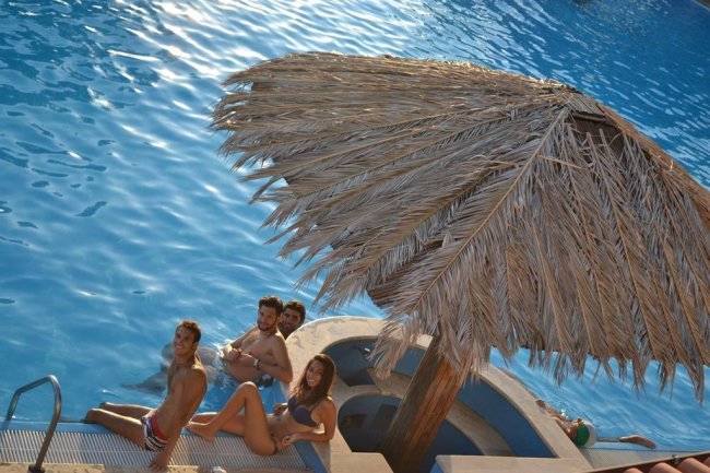 Persone in piscina sotto un ombrellone di paglia, acqua blu intorno.