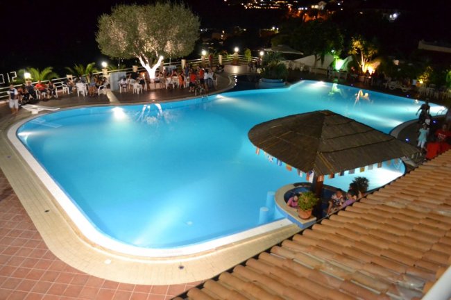 Veduta notturna di piscina illuminata circondata da persone e alberi.