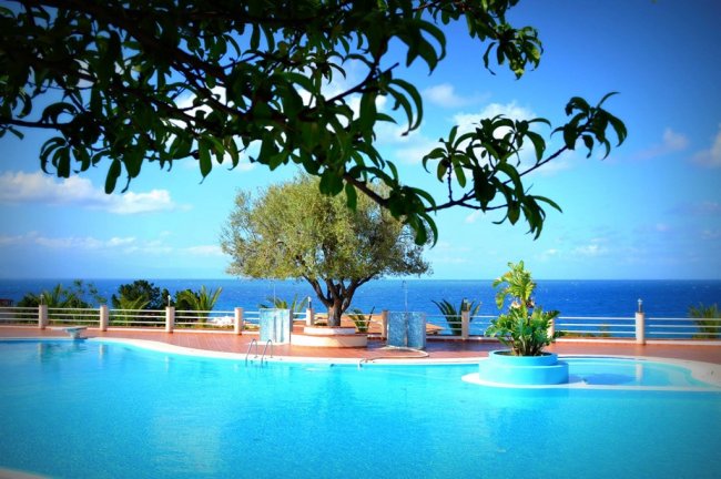 Piscina con vista mare, circondata da alberi e vegetazione.