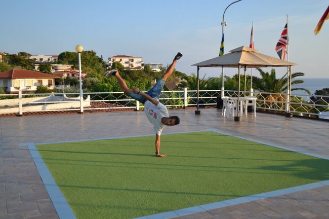 Una persona fa un'acrobazia su un tappeto verde, bandiere sventolano sullo sfondo.