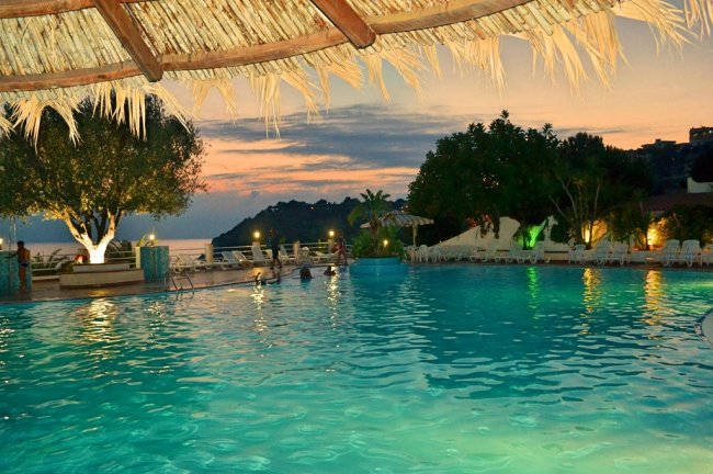 Piscina illuminata al tramonto con ombrellone di paglia sopra.