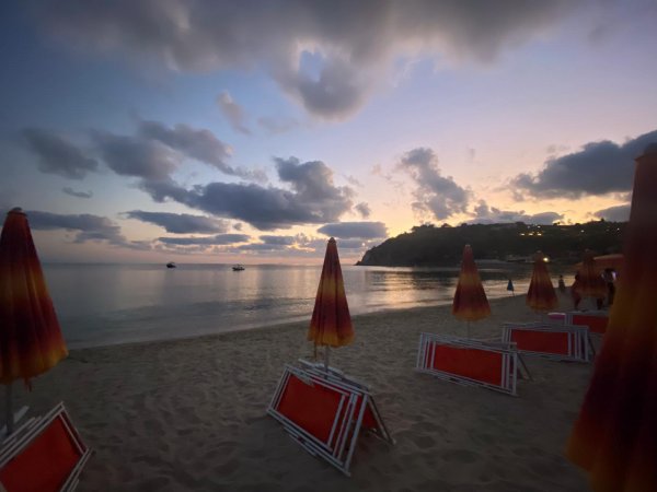 Spiaggia al tramonto con ombrelloni chiusi e cielo nuvoloso. Barche visibili a largo.