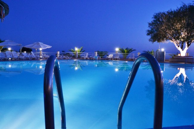 Piscina illuminata di sera con palme e albero.
