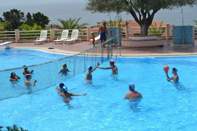 Persone giocano a pallanuoto in piscina all'aperto, con una rete e ombrelloni sullo sfondo.
