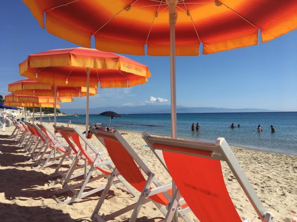 File di lettini con ombrelloni arancioni su spiaggia, persone in acqua calma sullo sfondo.
