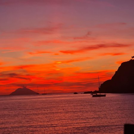 Tramonto infuocato su mare calmo con isola e barche a vela.