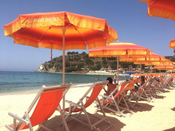 Spiaggia con sedie a sdraio arancioni sotto ombrelloni coordinati, mare e colline sullo sfondo.
