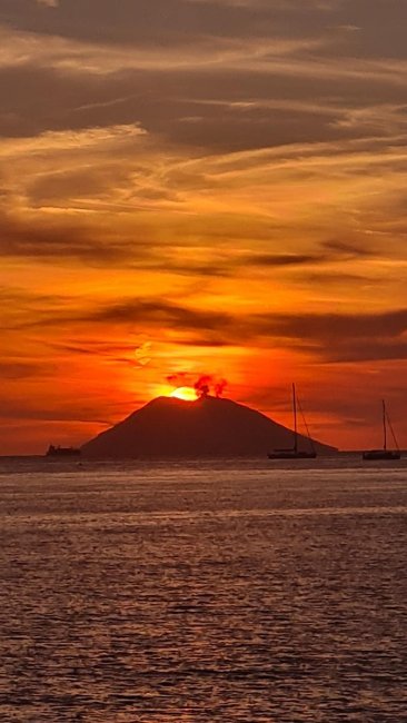 Sole al tramonto dietro un'isola con fumo, cielo rosso e barche a vela.