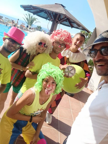 Sei persone vestite in modo colorato e festoso posano vicino a una piscina sotto un ombrellone.