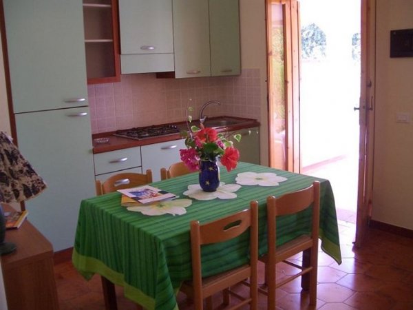 Cucina con tavolo coperto da tovaglia verde, fiori in vaso e porta aperta sul balcone.
