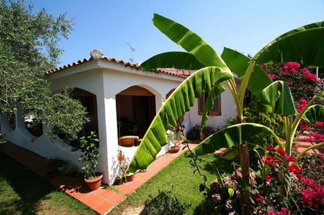 Casa mediterranea con giardino, grandi foglie di banano e fiori colorati.