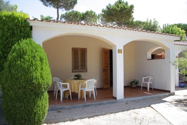 Veranda di una villetta con tavolo rotondo, sedie bianche e alberi sullo sfondo.