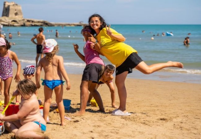 Persone che giocano e si divertono sulla spiaggia. Sullo sfondo, il mare e una torre.